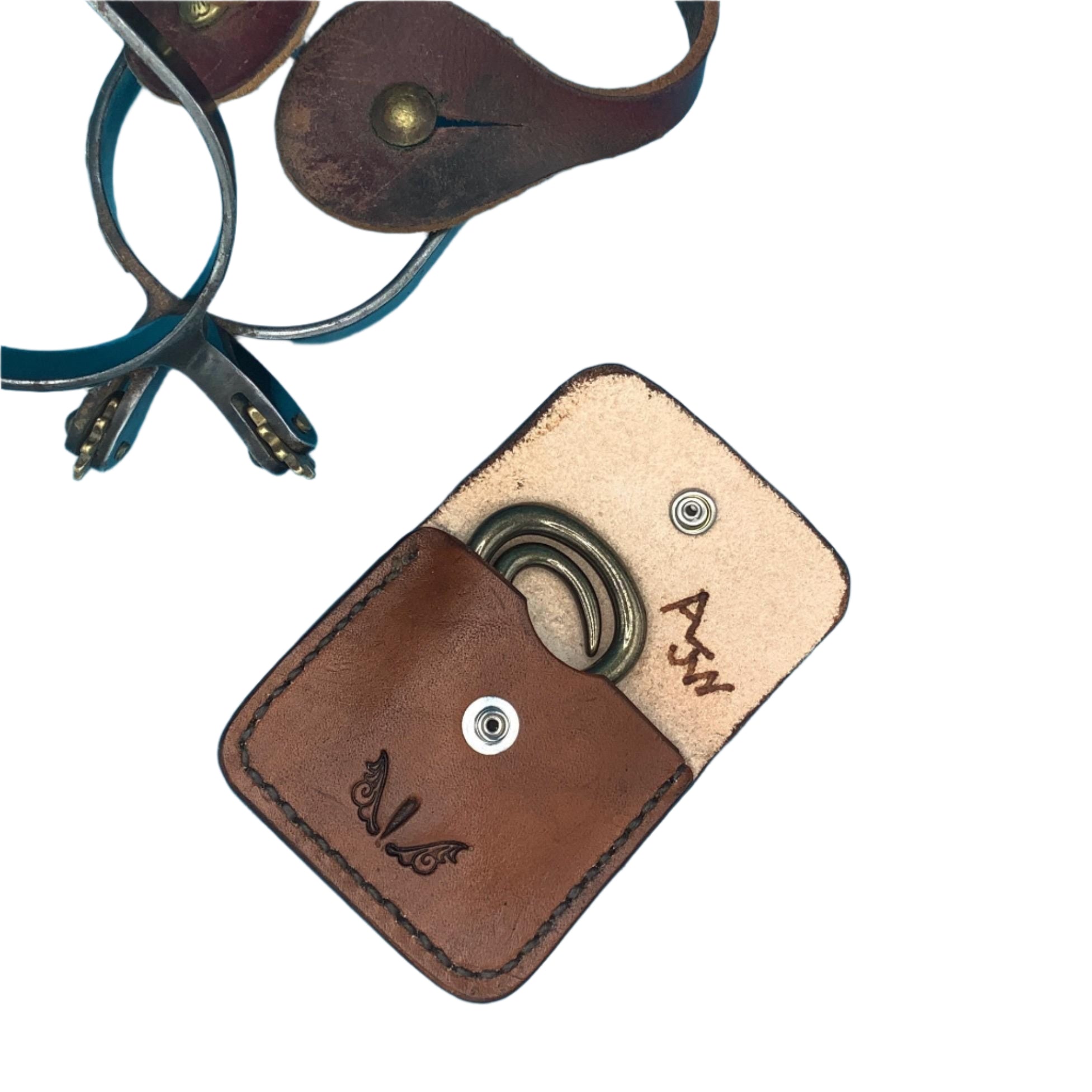A medium brown leather folding hoof pick  holder for a saddle billet with a folding hoof pick in it and spurs in the background.