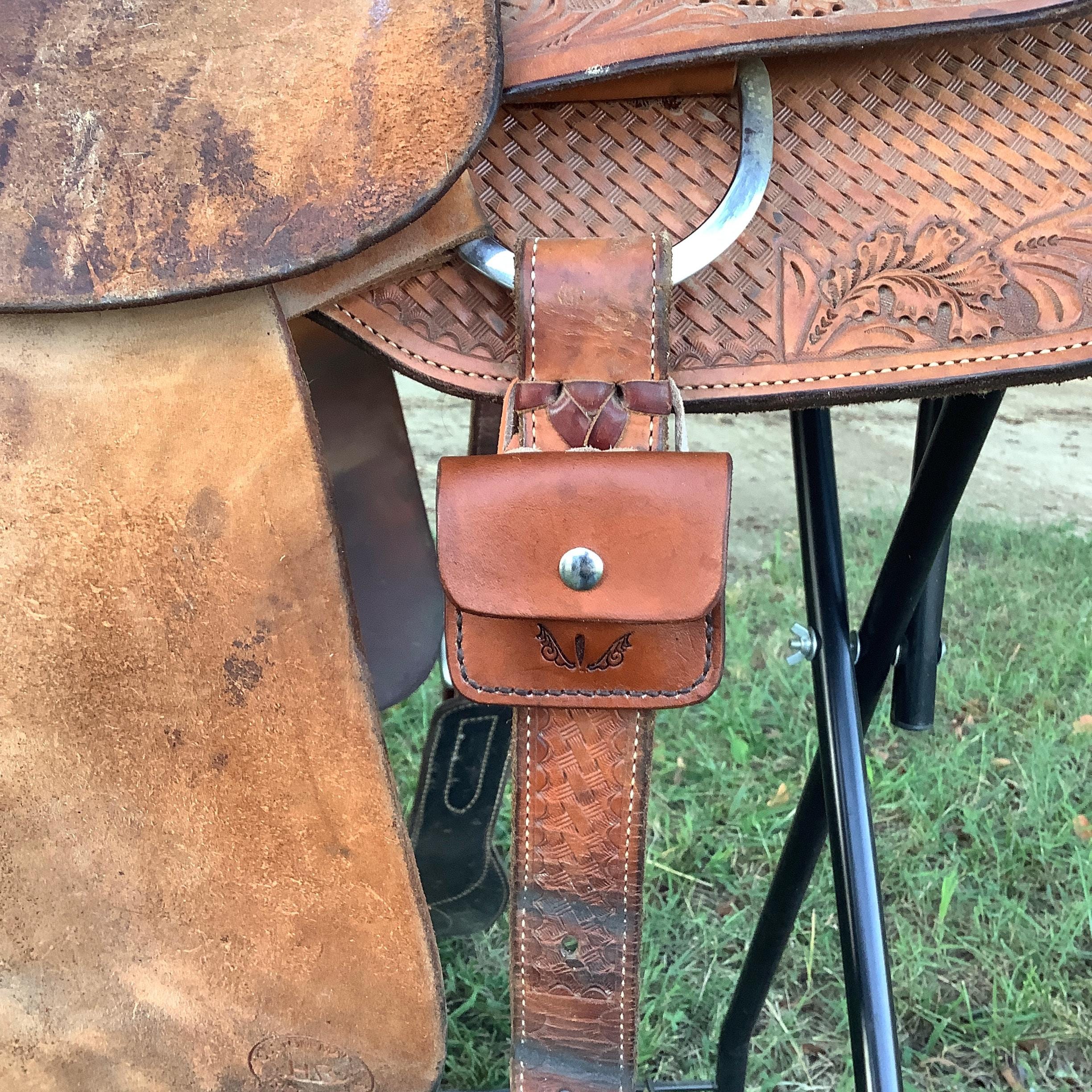 A leather folding hoof pick holder shown attached to the rear cinch of a saddle.